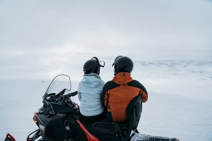 Två personer sitter på en skoter och blickar ut mot fjällen